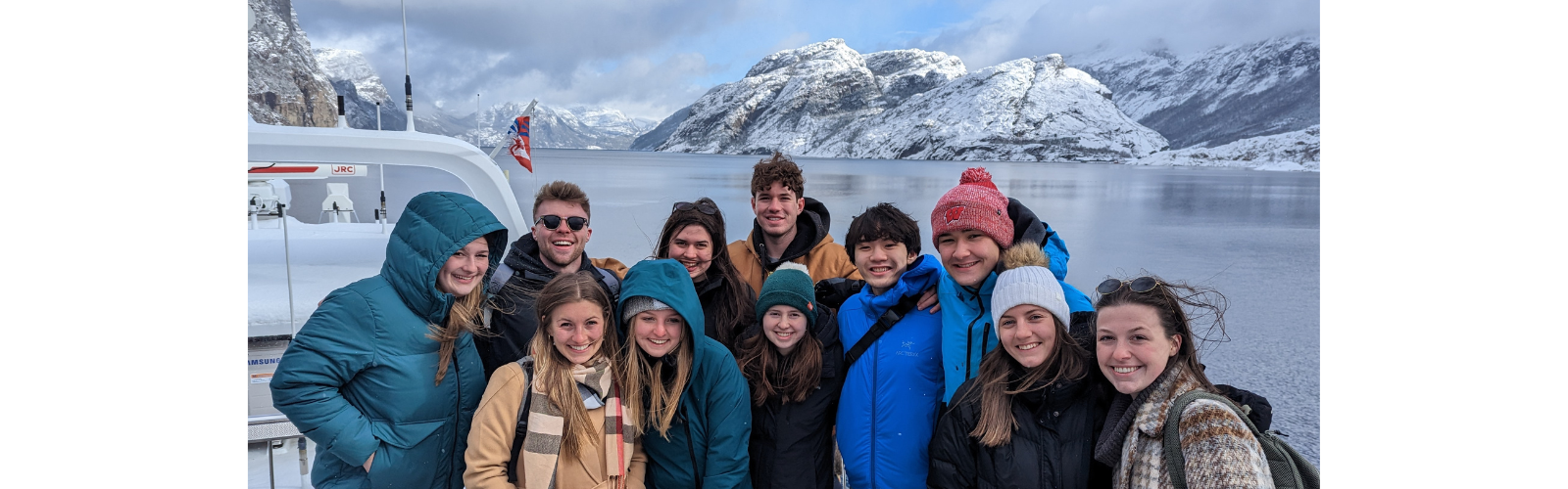 Students in warm clothes stand in front of a lake and several snowy mountains. 